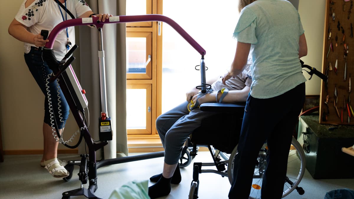 Kaksi hoitajaa on auttamassa pyörätuolissa istuvaa ihmistä ylös tuolista.