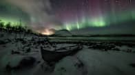 Photo of a snowy landscape at night with an old rowboat nestled by a lake, with the northern lights shimmering over a mountain in the night sky.