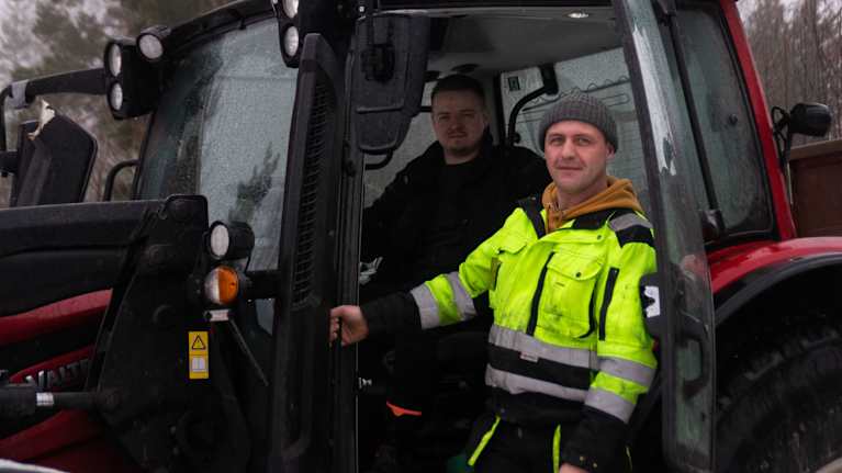 En man sitter och en står vid en traktor och ser mot kameran.