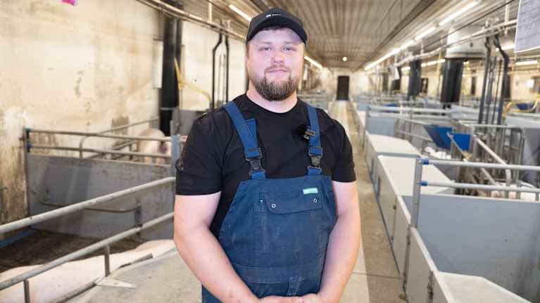 Personen står i en stor ladugård eller gård med flera bås omkring. Det ser ut som en arbetsplats för djurhållning eller jordbruk. Han bär arbetskläder inklusive en mörkblå hängselbyxor och en svart tröja.