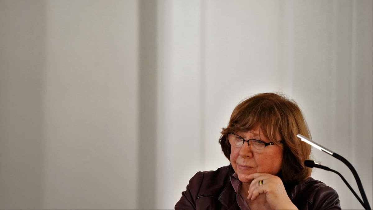 Silmälasipäinen Svetlana Aleksijevitš istuu kahden mikrofonin ääressä mietteliään näköisenä. Taustalla näkyy valkoista seinää.