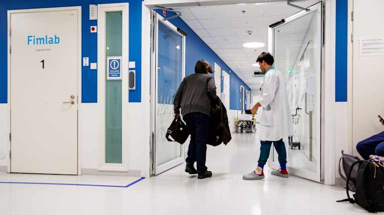 Doctor in white coat opens glass door for an older woman wearing grey top and dark pants in a brightly lit, white and blue medical facility.