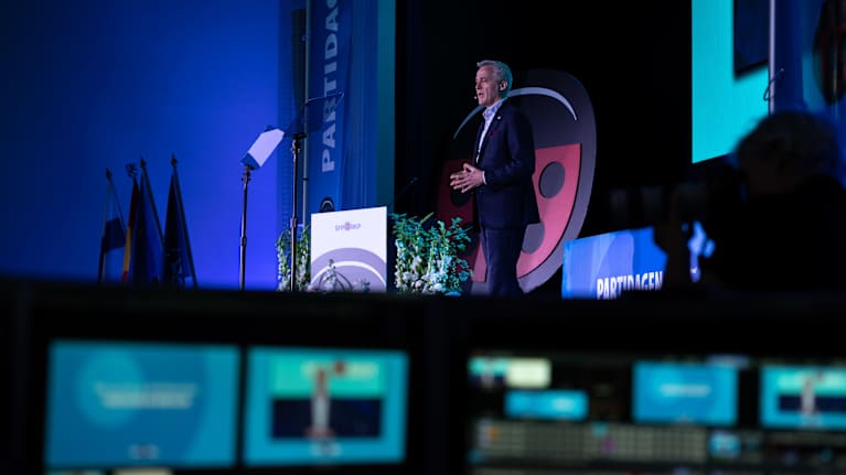 Bilden visar en person som talar vid ett talarpodium under ett partimöte eller kongress. I förgrunden syns kontrollbord med flera skärmar för teknikhantering av evenemanget.