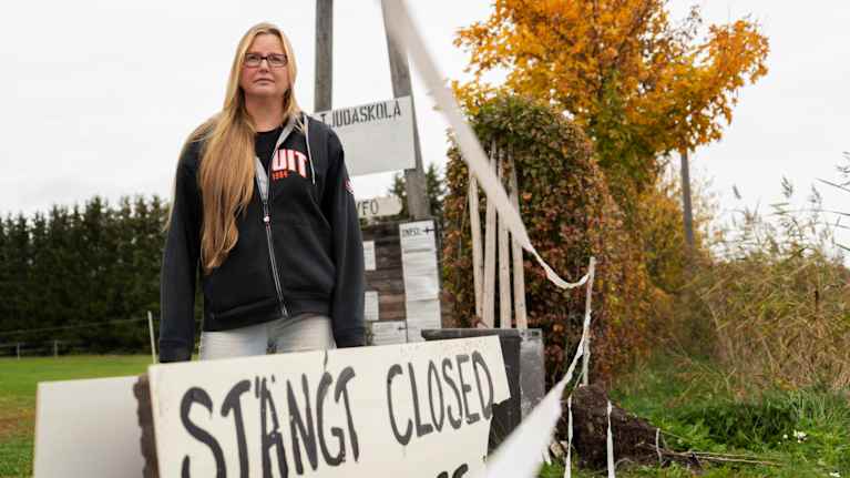 Kvinna står intill en infart. Vid infarten hänger ett vitt plakat där det står "STÄNGT CLOSED SULJETTU" med stora bokstäver.