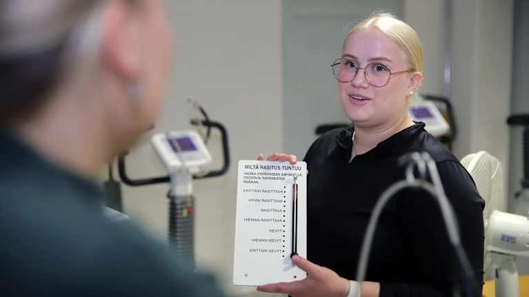 Opiskelija näyttää kuntopyörää polkevalle toiselle opiskelijalle rasitustaulukkoa Tanhuvaaran urheiluopiston testaustilassa.