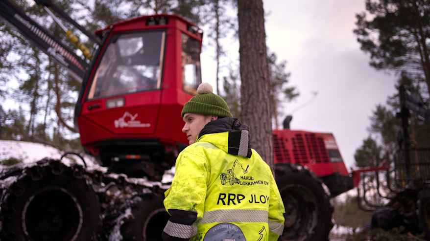 Nuori mies keltaisessa työtakissa ja vihreässä pipossa seisoo metsässä ja katsoo sivulleen, selin kameraan ja kädet taskussa. Taustalla näkyy metsäkone. 