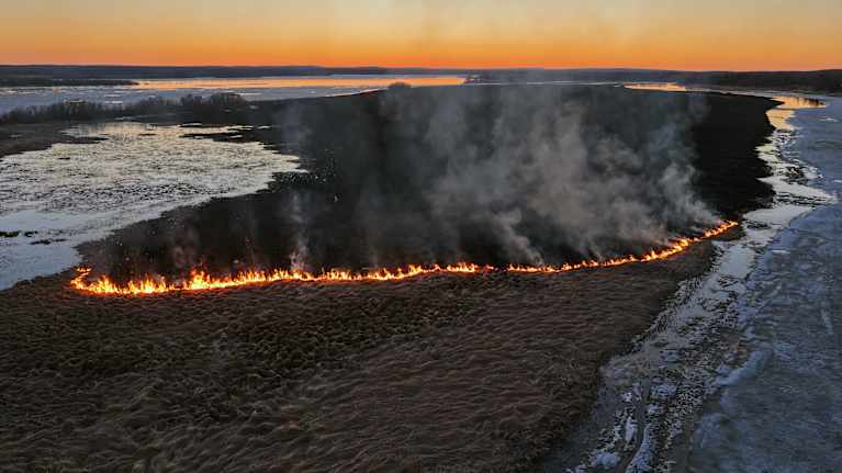 An aerial photo of a wildfire on an uninhabited grassy island. The evening sky is glowing orange and yellow and smoke is rising in the air.
