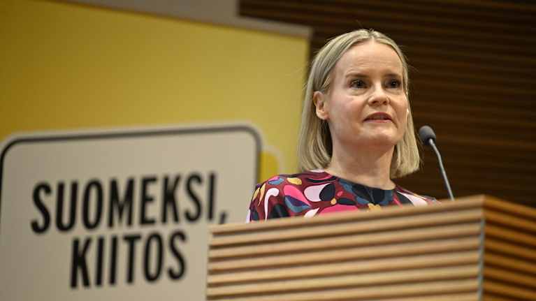 A blonde woman in a floral dress stands speaking at a podium in front of a sign saying "In Finnish, thanks" in Finnish.