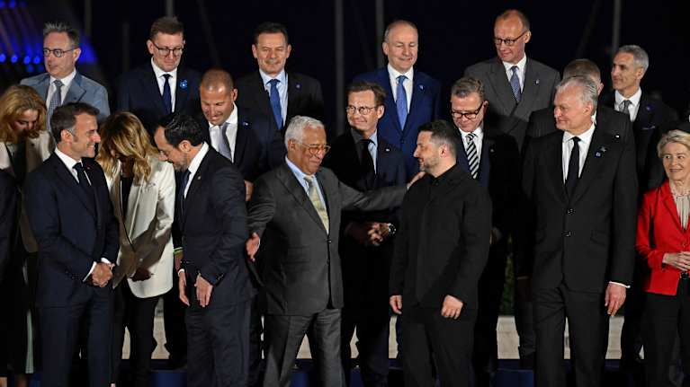 14 men and three women in suits stand in three rows against a dark background, some smiling, chatting and gesturing.
