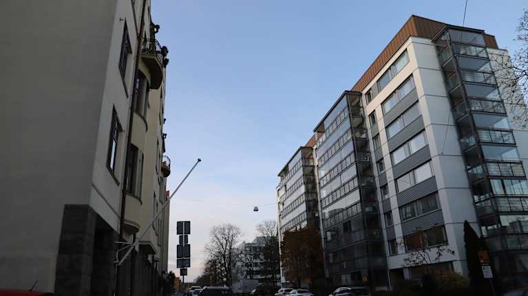 Kuva esittää kaupunkinäkymää kadulta. Kadun molemmin puolin on korkeita kerrostaloja. Vasemmalla puolella on vanhempia rakennuksia, joissa on pieniä parvekkeita. Oikealla puolella on moderneja, lasitetuin parvekkein varustettuja rakennuksia. Kadulla on parkkeerattuja autoja ja vähän liikennettä. Taivas on kirkas ja siinä näkyy auringonlaskun vaaleita sävyjä. Muutama ihminen kävelee kadulla kauempana. Ympäristö näyttää olevan urbaani ja rauhallinen.
