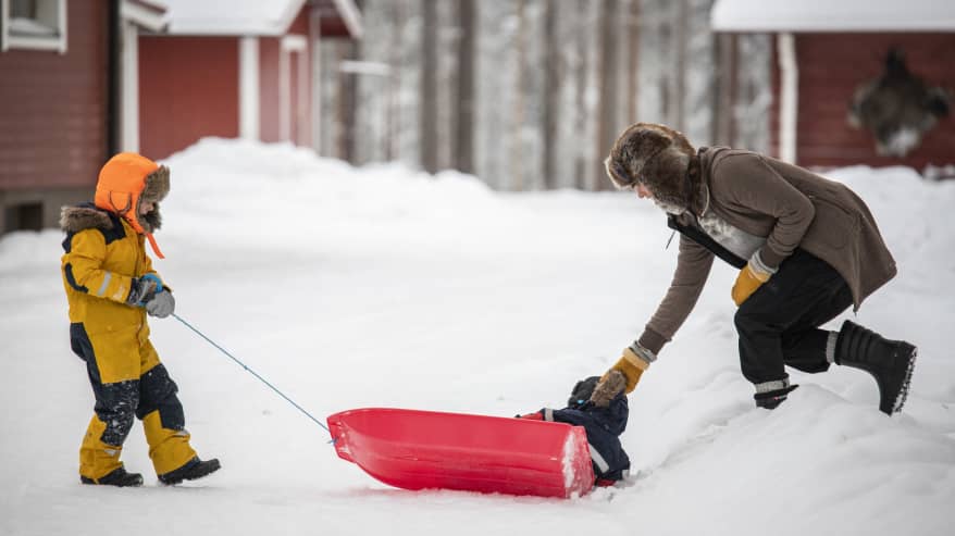 Äiti auttaa pulkasta kellahtanutta lastaan, toinen lapsi vetää pulkkaa.