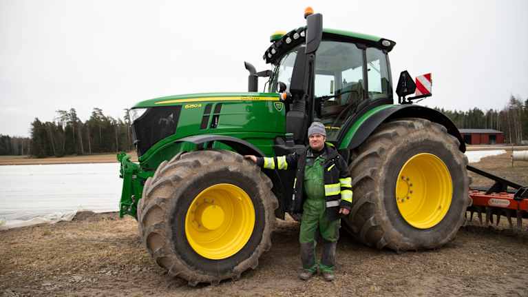 En person står vid sidan av en stor grön traktor, med jordbruksmark i bakgrunden.