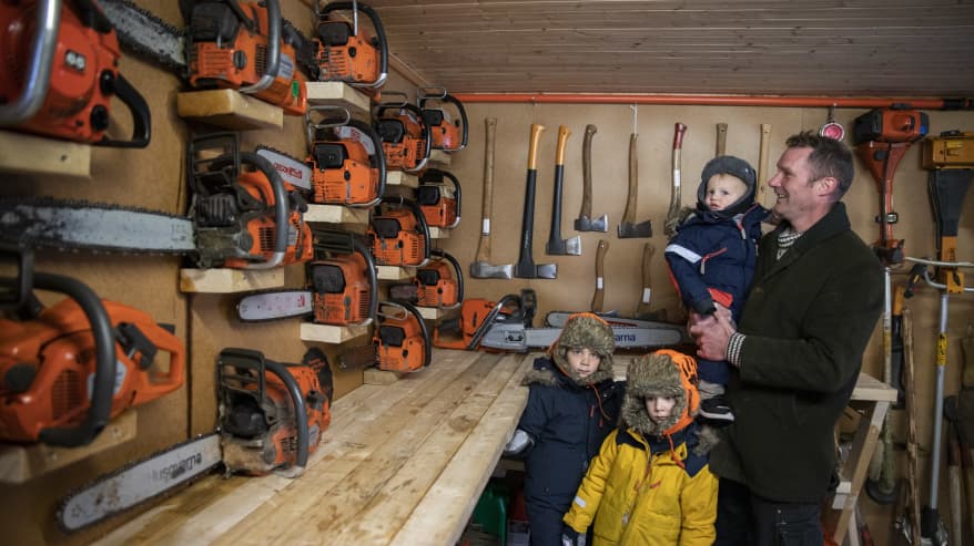 Isä ja kolme pientä poikaa katselevat seinälle aseteltuja moottorisahoja työkaluvarastossa.