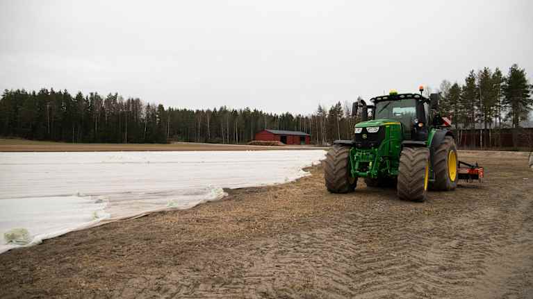 En traktor står parkerad vid kanten av ett fält täckt med vit plast, med en bakgrund av en skog och en röd byggnad.