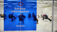 People sitting in a large white waiting room, behind a translucent blue sign that reads "Covid vaccine point Hansa Kymsote" in Finnish.
