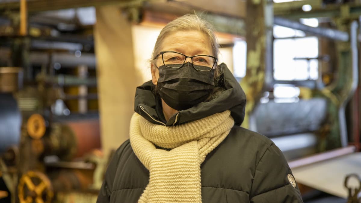 Tarja Peltola-Welin seisoo vanhassa tehdashallissa maski kasvoillaan ja talvitakki yllään. Katsoo kohti kameraa.