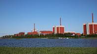 A series of large dark red and grey buildings along a shore, seen from across the water with reeds in foreground. 