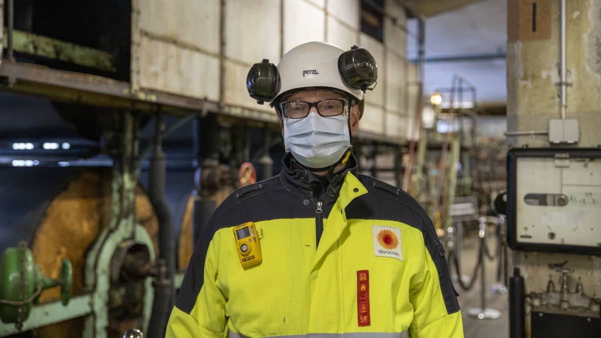 Antti Veitola seisoo vanhassa tehdashallissa työtakki- ja kypärä päässään ja maski kasvoillaan