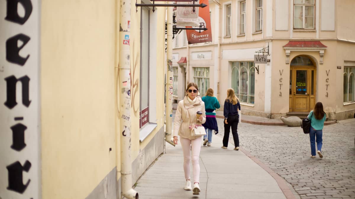 Nainen aurinkolaseissa, vaaleassa takissa ja vaaleanpunaisissa housuissa kävelee vanhankaupungin katuja.