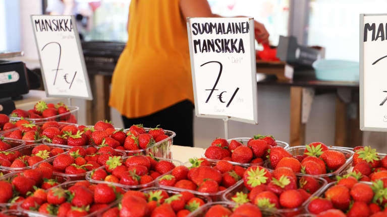 Jordgubbor i rivor på ett torgstånd. I bakgrunden en torgförsäljare i en apelsinfärgad skjorta. 