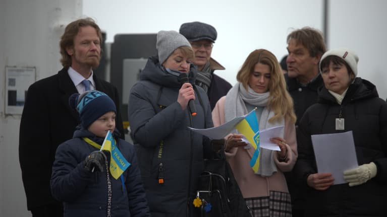 Joukko suomalaisia ja ukrainalaisia. Nainen puhuu mikrofoniin. Henkilöiden kädessä on Ukrainan lippuja.