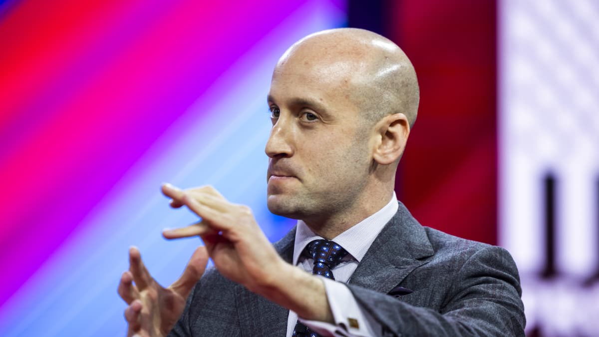 Stephen Miller elehtii käsillään, tiukka ilme, värikäs tausta. 