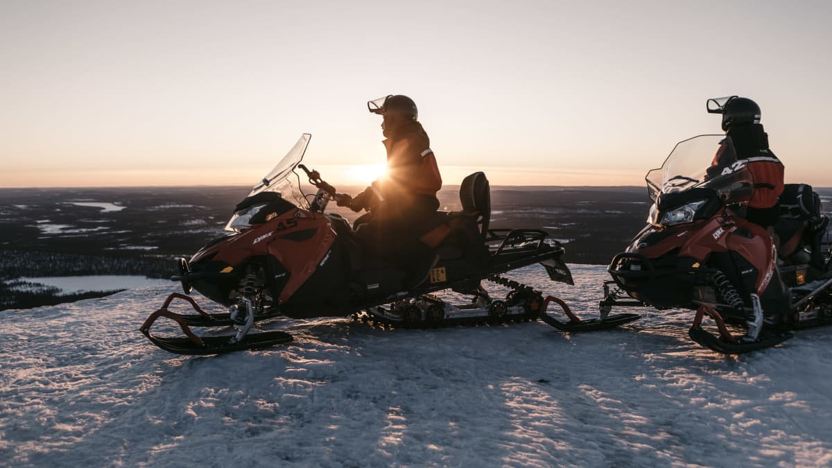 Photo shows two people on snowmobiles, stopping to look at a sunset.