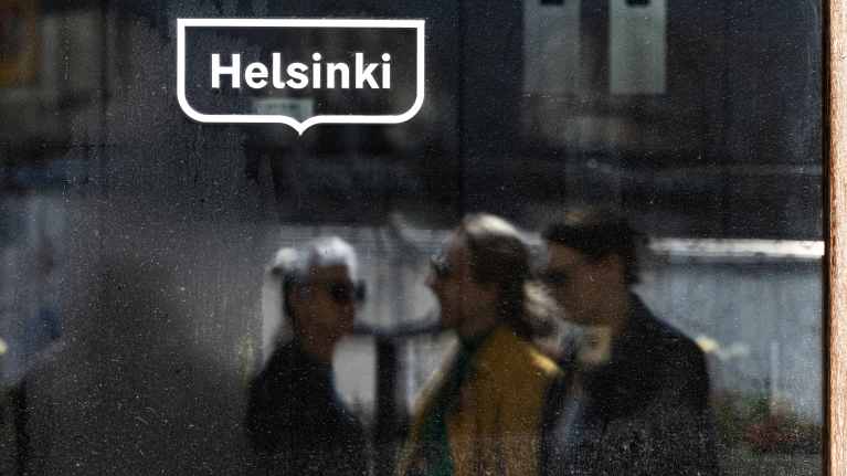 Ikkunaan liimattu Helsingin kaupungin logo ja ikkunaan heijastuu ohi kulkevia henkilöitä.