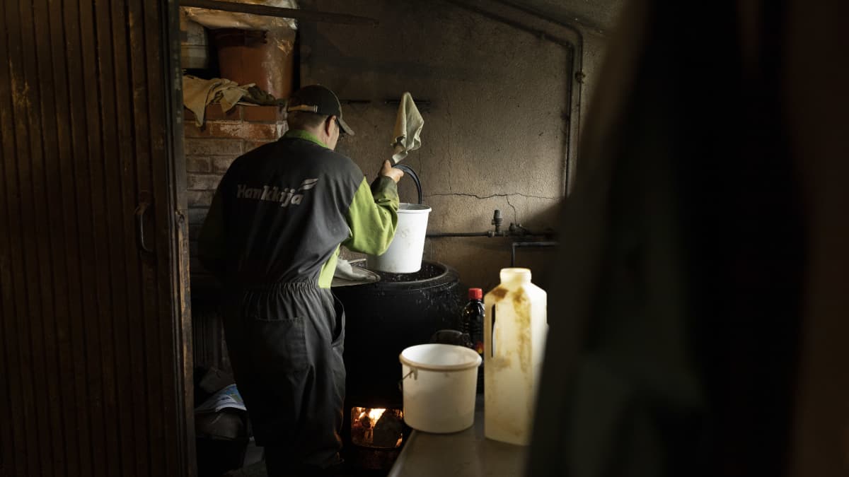 Timo Liimatainen nostaa karjakeittiössä ämpärillä kuumaa vettä padasta, jonka alla palaa tuli. 