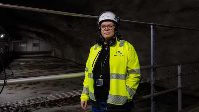 Mirva Levomäki, en person med skyddshjälm och reflexjacka står i vid bassänger i ett vattenreningsverk med betongväggar.