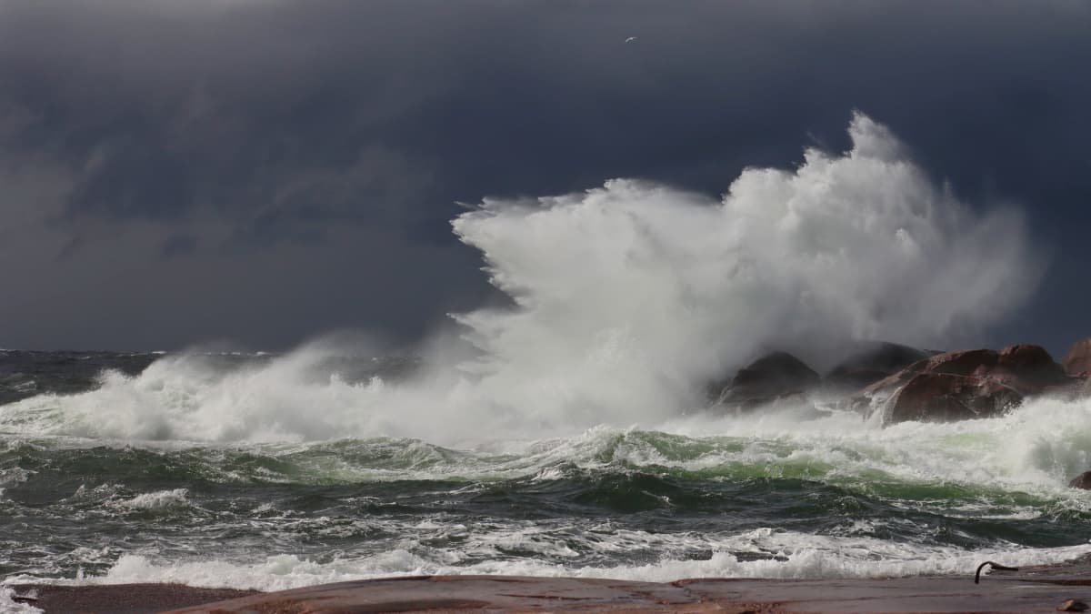 Aalto lennättää merivettä useiden metrien korkeuteen sen osuessa rantakivikkoon. Taustalla on tumma pilvimassa.