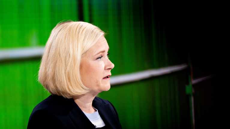 Profile shot of a blonde woman in a dark suit speaking against a green backdrop in a TV studio. 