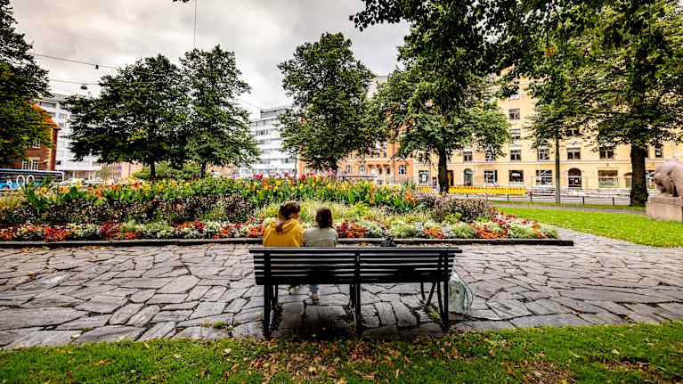 Two people sit on a park bench in front of a colorful flowerbed.