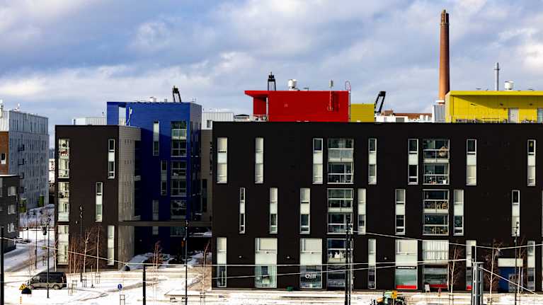 A winter cityscape. In the foreground are modern, multi-storey buildings with largely dark facades. On the upper floors, however, there are brightly coloured elements such as blue and red.