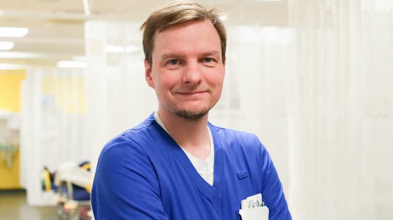 Personporträtt på ung, manlig sjukskötare klädd i blå arbetskläder. Han ler och tittar in i kameran. I bakgrunden vårdavdelning med platser med fördragna skynken.