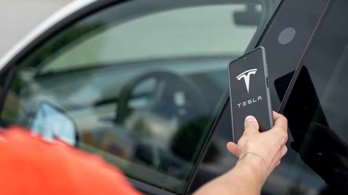Henkilö pitää puhelinta, jossa näkyy Teslan logo, auton oven vieressä.