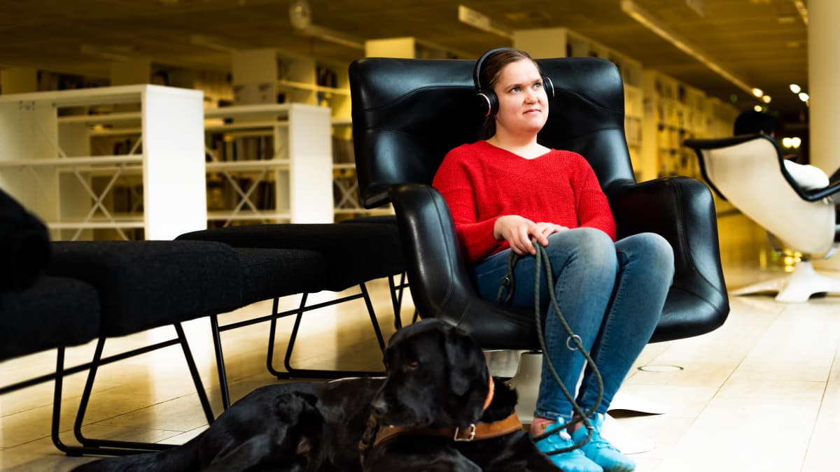 Susanna Halme istuu kirjastossa nojatuolissa ja kuuntelee äänikirjaa. Vierellä koira.