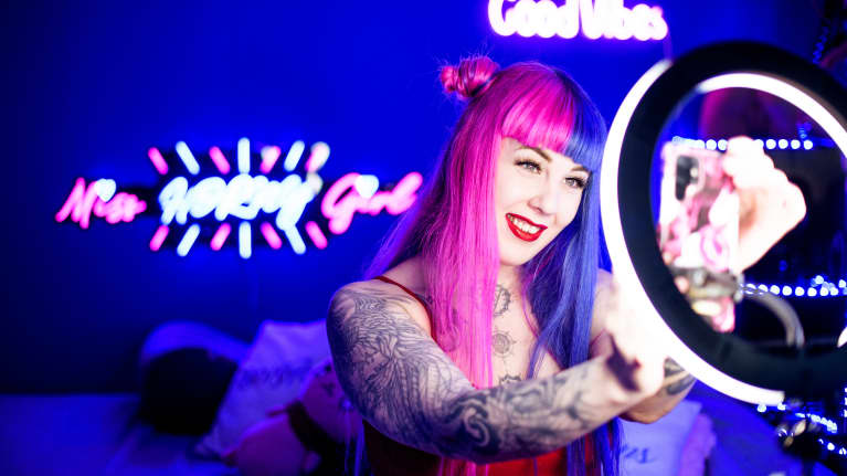 Nainen poseeraa kännykkäkameralleen huoneessa, jossa on neonvaloja ja kuvausvalo. 