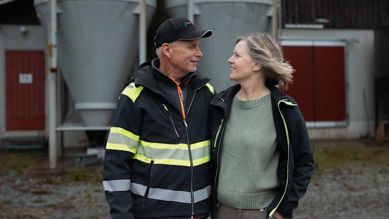 Två vuxna personer står utomhus framför två stora silos och en byggnad med röda dörrar.