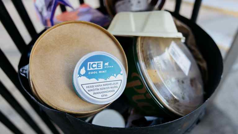 Kuvassa on roskakori, johon on kerätty erilaisia roskia, kuten jäätelökartonki ja muovipakkaus. Erottuvasti näkyy Ice Cool Mint -purkkakuorinen pakkaus, joka on asetettu roskien päälle.