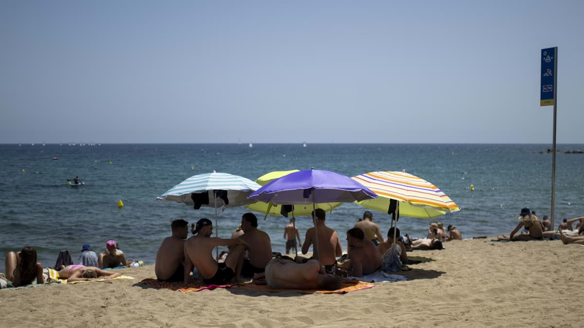 Ihmisiä rannalla. Suuri joukko istuu aurinkovarjojen alla tiiviissä ryhmässä.