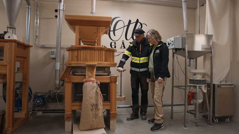 Två personer står vid en kvarnmaskin i ett rum med industrimaskiner; på väggen syns texten "Otto Gård".