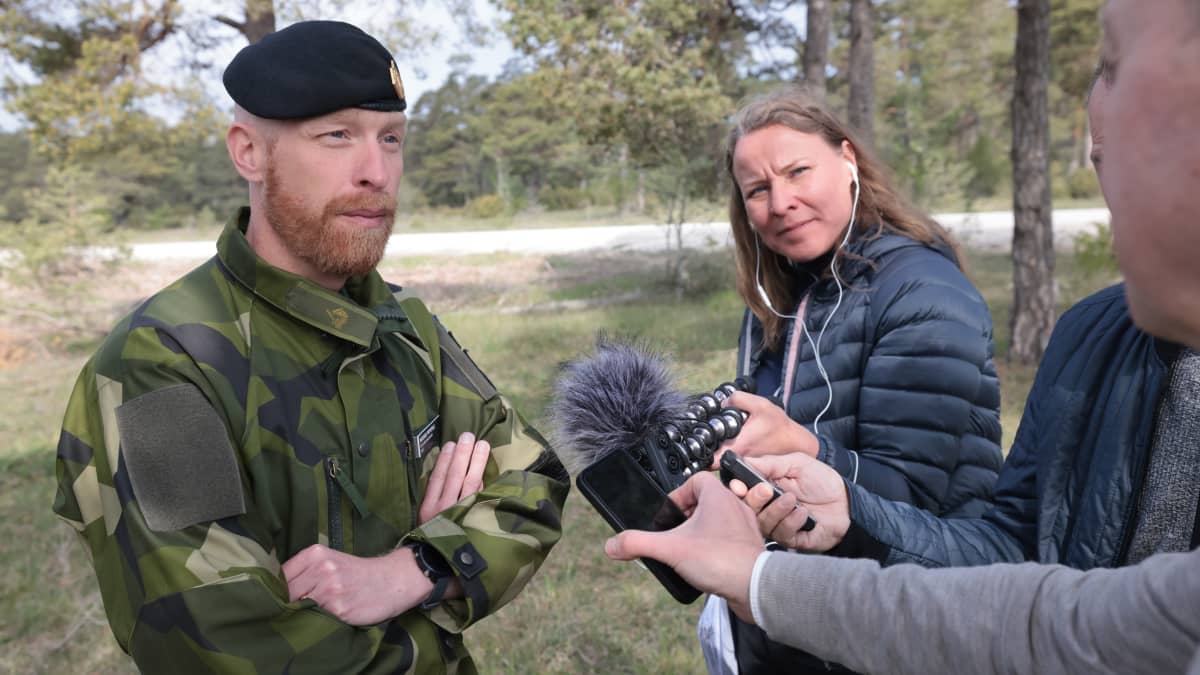 Armeija-asuinen mies haastateltavana, haastattelijasta näkyy vain hieman sivuprofiilia. Taustalla nainen seisoo ja katsoo tilannetta.