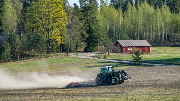 En grön traktor harvar jorden en vårdag där löven spricker ut i träden och en röd lada lyser i solen.