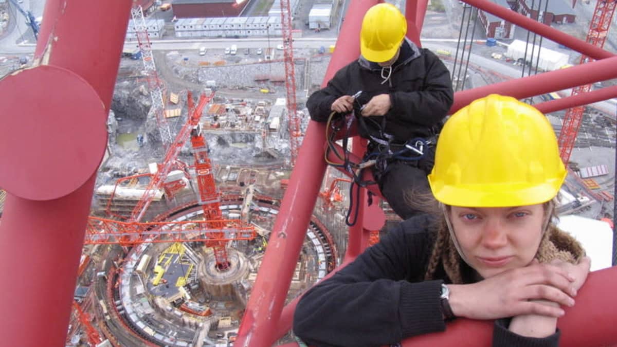 Nainen keltainen kypärä päässään katsoo kameraan korkealla nosturin nokassa Olkiluoto 3:n työmaalla, taustalla toinen henkilö.