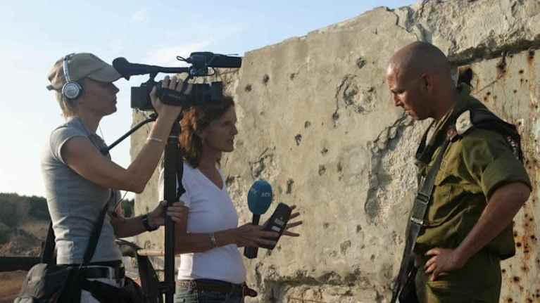 TV-fotograf med keps och beige t-skjorta och journalist med vit t-skjorta intervjuar en israelisk soldat med gröna kläder och ett gevär i handen.