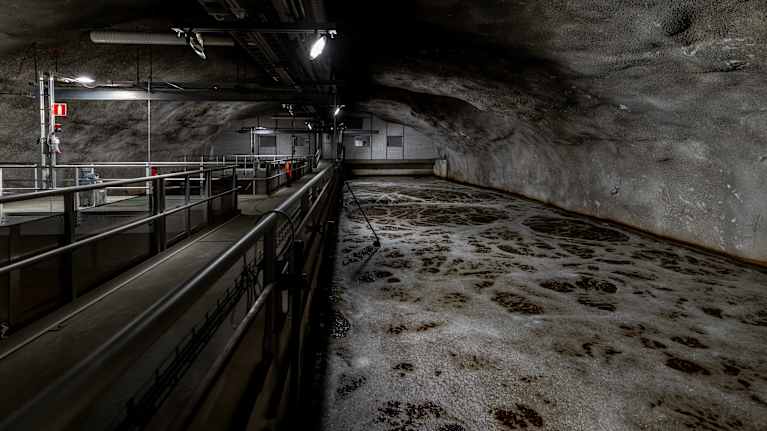 En underjordisk anläggning för avloppsvattenrening med gångbro och stora bassänger täckta av skum och vatten.