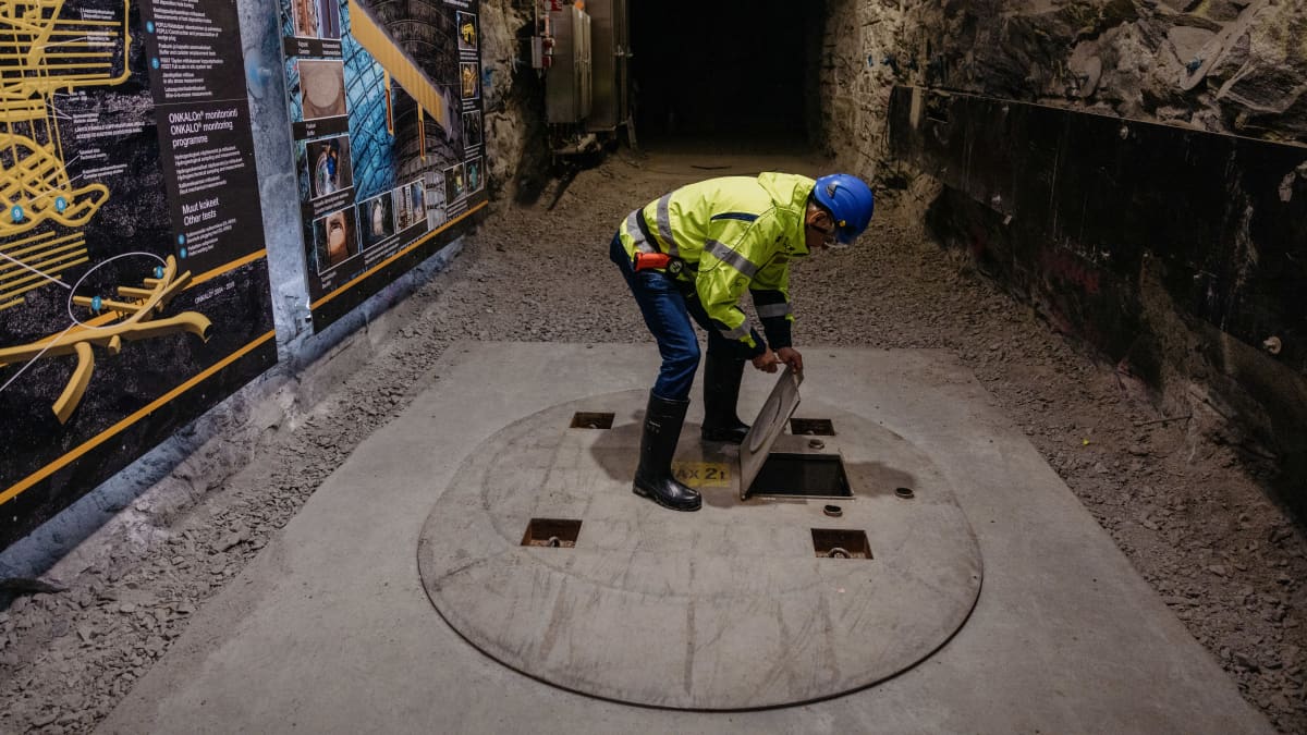Man wearing bright yellow jacket and blue helmet, lifting a metal door on the floor of a long, rocky tunnel.