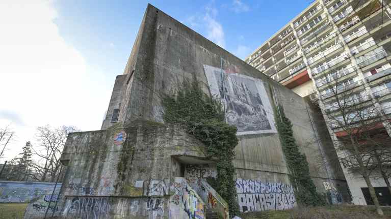 Korkea betoninen, likaantunut ja alareunastaan graffitien peitossa oleva karu bunkkeri tulee ulos noin 1970-luvun kerrostalon sisältä. Vaalea kerrostalo parvekkeineen on rakennettu bunkkerin ympärille.