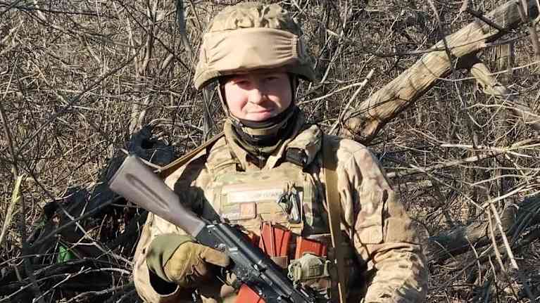 Keskustapoliitikko Ville Mykkänen seisoo täydessä taisteluvarustuksessa rynnäkkökiväärin kanssa ja katsoo kohti kameraa neutraali ilme kasvoillaan.